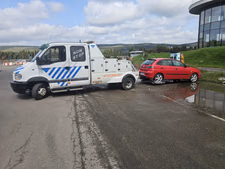 Tractare autoturism mic — un hatchback roșu (Seat Ibiza), defect, ridicat pe platforma cu cârlig a mașinii de tractare, lângă o clădire modernă cu fațadă vitrată.