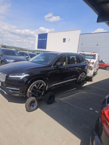 Tractare autoturism — un SUV Volvo XC90 negru, cu roata față ridicată pe un cărucior/osie auxiliară de tractare, remorcat lângă un showroom Volvo. Intervenție de asistență rutieră pentru mașină cu defecțiune la roată/suspensie.