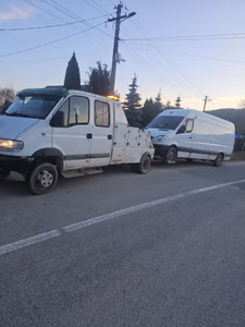 Tractare la asfințit, pe un drum de țară. Dubă albă de marfă (Mercedes Sprinter), cu avarie frontală, remorcată cu platforma cu cârlig