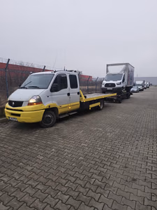 Zi cu ceață, curte industrială. Un camion tip Ford Transit cu cutie de marfă este încărcat integral pe platforma galbenă a mașinii de tractare, lângă alte autoutilitare aliniate.
